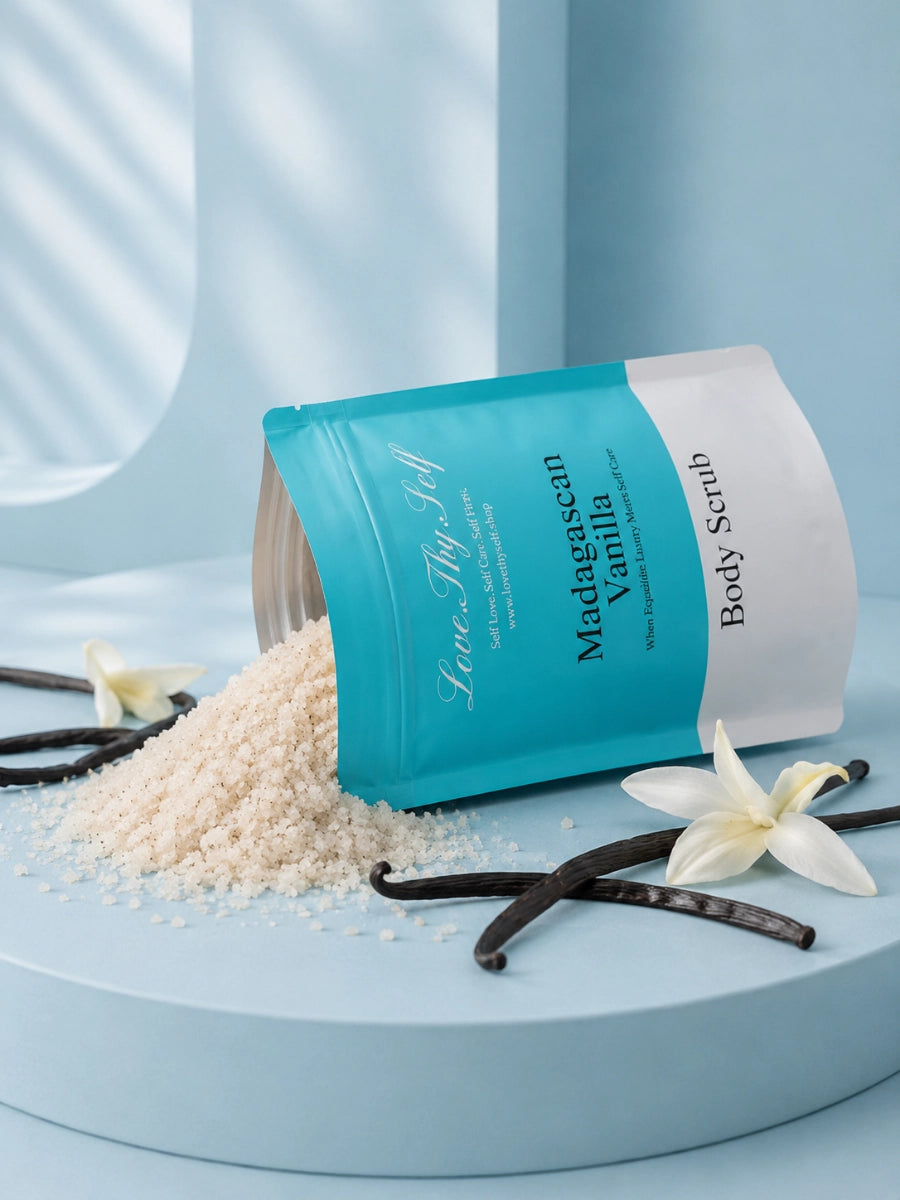 LoveThySelf Madagascan Vanilla Body Scrub with exfoliating scrub spilling from an open pouch, styled with vanilla pods and white flowers.